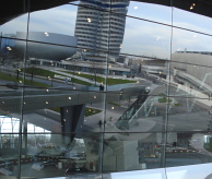 De afhaalzone van de BMW Welt, gezien door het raam met daarin weerspiegeld: het BMW Museum, het hoofdkantoor en de fabriekshal (v.l.n.r.). Afbeelding: Carina de Jonge